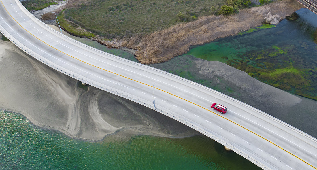 Aerial Drone Photo of SUV with Surf Board on roof crossing coastal bridge