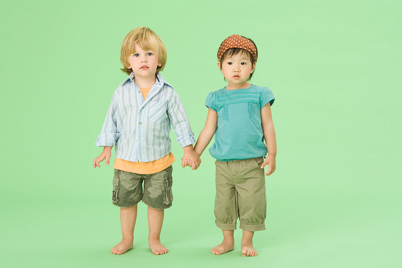 Photograph of a caucasian toddler boy and an asian toddler girl on green seamless paper holding hands