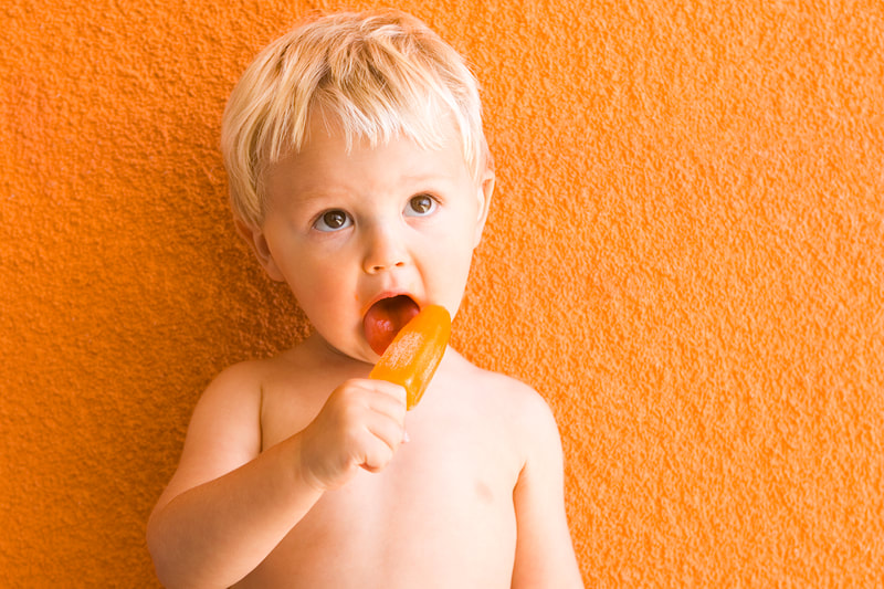 Photograph of a caucasian baby boy eating an orange popsicle against an orange stucco background